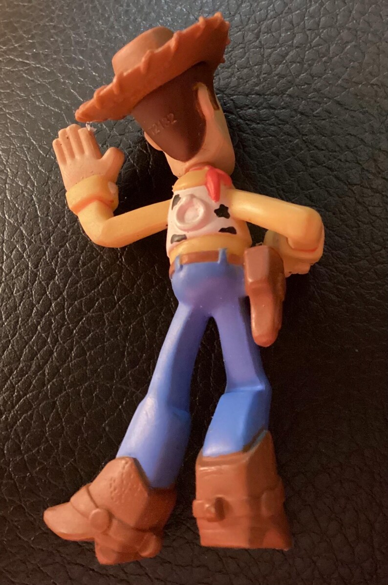 Pu&ograve; includere: Una piccola statuetta in plastica di un personaggio cowboy che indossa un cappello marrone, una camicia gialla con macchie nere, un fazzoletto rosso, pantaloni blu e stivali marroni. La figura ha una fondina sul fianco sinistro e un anello sul retro.