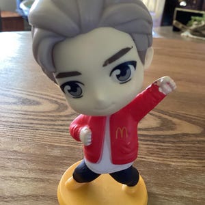 May include: A figurine of a cartoon character with gray hair, wearing a red jacket with the McDonald's logo, black pants, and yellow shoes. The figure is standing on a yellow base.
