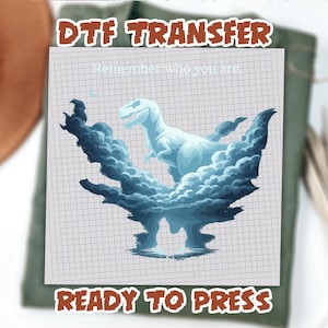 May include: DTF transfer design featuring a light blue Tyrannosaurus Rex illustration surrounded by dark blue clouds. The text "Remember who you are" is above the dinosaur, and "READY TO PRESS" is below. The design is on a grid background.