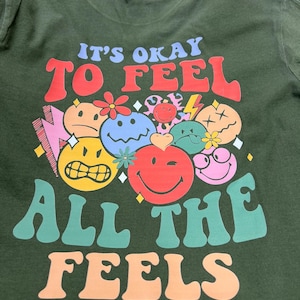 May include: Dark green t-shirt with the text "IT'S OKAY TO FEEL ALL THE FEELS" in colorful, retro-style lettering. The shirt features a collection of smiling faces and other graphic elements.