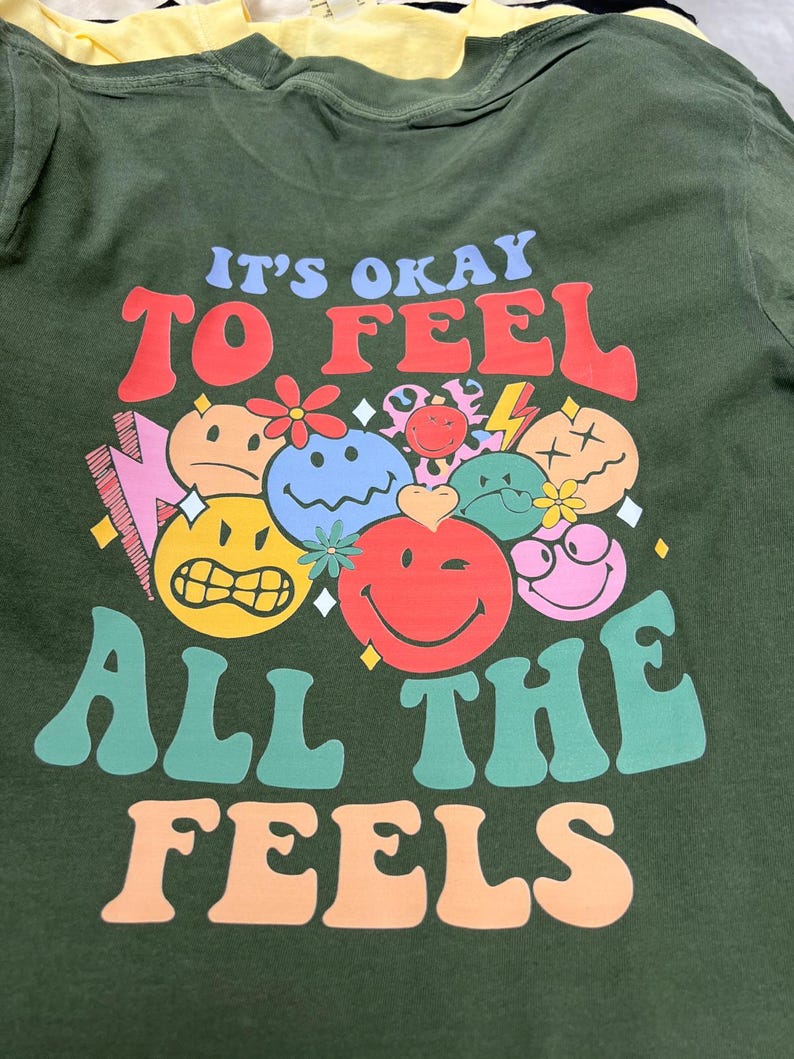 May include: Dark green t-shirt with the text "IT'S OKAY TO FEEL ALL THE FEELS" in colorful, retro-style lettering. The shirt features a collection of smiling faces and other graphic elements.