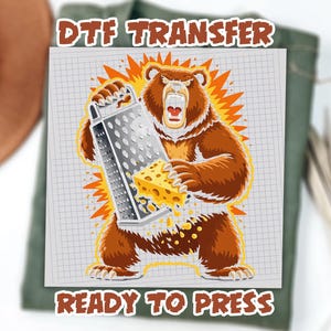May include: DTF Transfer design featuring a cartoon bear grating cheese with a cheese grater. The bear is brown with an open mouth and is surrounded by an orange burst. The words "DTF TRANSFER" and "READY TO PRESS" are in the image.