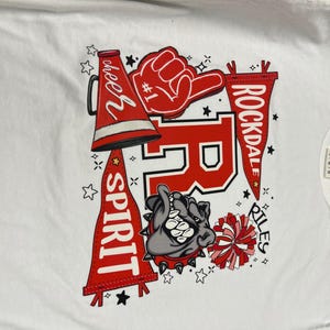 May include: White t-shirt featuring a Rockdale Riles design. The design includes a red megaphone, a foam hand, a bulldog, and pennants with the words "Cheer Spirit" and "Rockdale Riles".