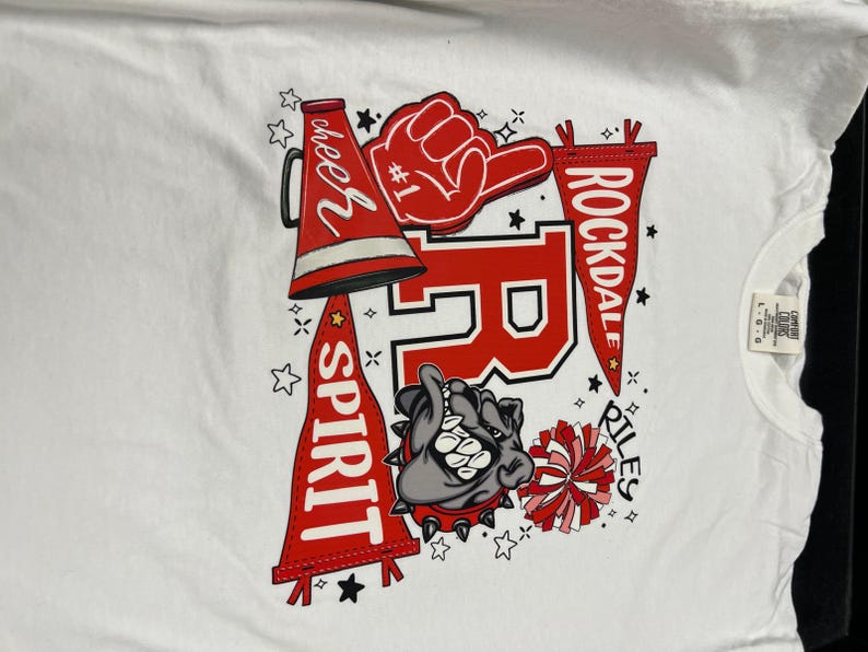 May include: White t-shirt featuring a Rockdale Riles design. The design includes a red megaphone, a foam hand, a bulldog, and pennants with the words "Cheer Spirit" and "Rockdale Riles".