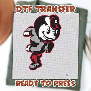 May include: A DTF transfer featuring a cartoon character in black, gray, and red, wearing a cap. The character is running with a determined expression. The text "DTF TRANSFER" and "READY TO PRESS" are displayed in a brown, outlined font.