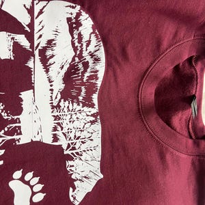 May include: Burgundy sweatshirt featuring a white graphic of a bear's head and paw print. The bear design incorporates a forest scene. The sweatshirt has a crew neck and is made of a soft material.