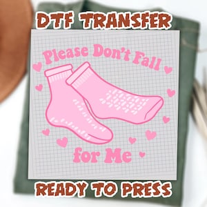 May include: A pink DTF (Direct to Film) transfer featuring two pink socks with the text "Please Don't Fall for Me" and hearts. The words "Ready to Press" are also included. The design is on a grid-lined paper.