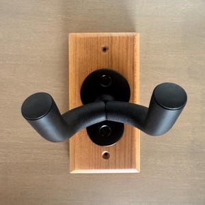 May include: A wall-mounted guitar hanger featuring a rectangular light brown wooden base with two pre-drilled screw holes. Attached to the center is a black metal bracket supporting two curved, padded black arms designed to hold a guitar neck.