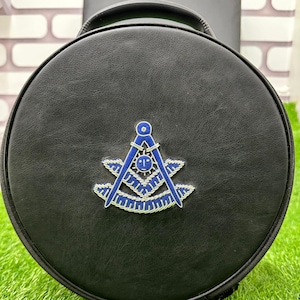 Op de afbeelding: Zwarte, ronde tas met een handvat. Het heeft een blauw en zilver embleem met een kompas en een vierkant. De tas ligt op kunstgras.