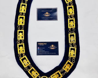 Op de afbeelding: Een donkerblauwe fluwelen kraag met goudkleurige rechthoekige schakels en stervormige accenten. De schakels hebben kleine figuren. Twee visitekaartjes zijn op de kraag geplaatst. De tekst op de kaarten is wazig.