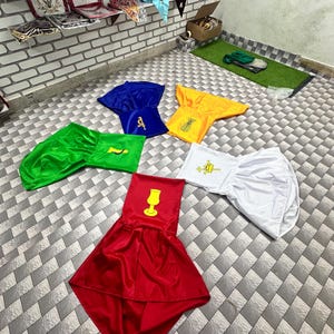 May include: A collection of brightly coloured ceremonial robes in red, blue, green, yellow, and white. Some robes have gold embroidered symbols. The garments are arranged on a tiled floor, with more items visible on shelves in the background.