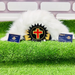 May include: A white, fluffy ceremonial hat with a black and gold emblem featuring a red cross. The hat rests on green artificial grass, with two small business cards on either side.