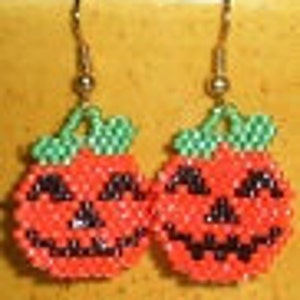 Puede incluir: Un par de pendientes de calabaza naranja y verde con rasgos faciales negros. Los pendientes están hechos de cuentas y tienen ganchos dorados.