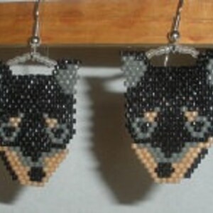 May include: A pair of beaded earrings shaped like a dog's head. The earrings feature a black body, gray ears, and a tan muzzle. The earrings are hanging from silver-colored ear wires.