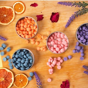 Puede incluir: Vista aérea de una tabla de madera con cuatro pequeños recipientes metálicos llenos de ceras aromáticas de colores. Las ceras son naranjas, rosas, azules y moradas. Rodajas de naranja secas, pétalos de rosa y ramitas de lavanda dispersas.