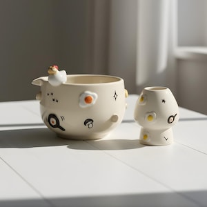 May include: A cream-colored ceramic bowl and vase set. The bowl features a chicken figurine, fried egg, and pan illustrations. The vase is mushroom-shaped with fried egg and star designs. Both are on a white surface.