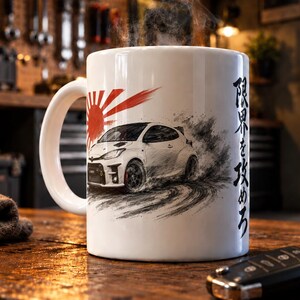 May include: A white ceramic mug featuring a graphic of a white hatchback car drifting on a track, a red sun emblem, and Japanese text. Steam rises from the top of the mug, which is positioned on a wooden surface next to a black car key fob.