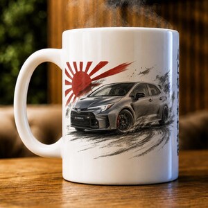 May include: A white ceramic mug featuring a graphic of a grey Toyota GR Corolla car drifting on a track. Behind the car is a red sun design with radiating rays. Japanese characters are printed vertically on the side of the mug near the handle.