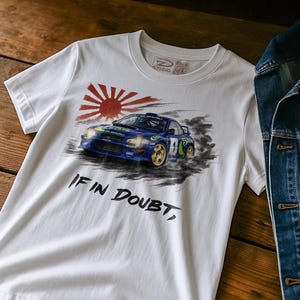 Może przedstawiać: Biała koszulka z ilustracją niebieskiego samochodu rajdowego Subaru i motywem wschodzącego słońca. Koszulka ma napis "IF IN DOUBT," czarnym pismem. Kurtka dżinsowa jest częściowo widoczna po prawej stronie.