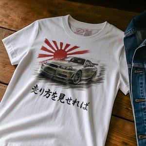 May include: White t-shirt featuring a car graphic with a rising sun design. The graphic includes a silver car and Japanese text. The shirt has a crew neck and short sleeves. A denim jacket is partially visible in the background.