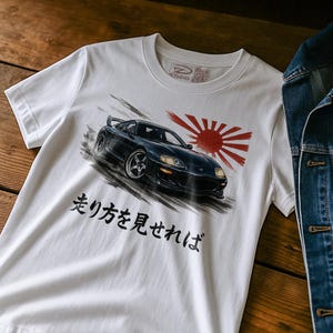 Könnte beinhalten: Weißes T-Shirt mit einer schwarzen Sportwagen-Grafik, einem roten Sonnenmotiv und japanischem Text. Das Auto wird in Bewegung mit Bewegungsstrichen dargestellt. Das T-Shirt hat einen Rundhalsausschnitt und kurze Ärmel.