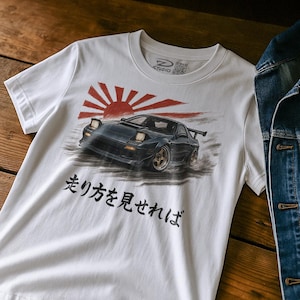 Könnte beinhalten: Weißes T-Shirt mit einer Grafik eines schwarzen Sportwagens und einer rot-weißen aufgehenden Sonne. Japanischer Text ist unter dem Auto aufgedruckt. Das T-Shirt hat einen Rundhalsausschnitt und kurze Ärmel.
