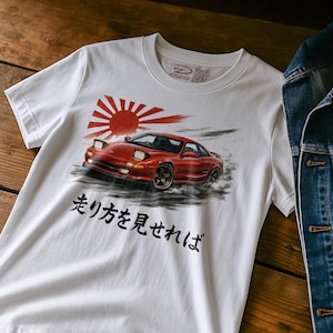 Könnte beinhalten: Weißes T-Shirt mit einer Grafik eines roten Sportwagens und einem Sonnenaufgangsdesign. Das Shirt hat japanischen Text und ein kleines Logo am Kragen. Eine Jeansjacke ist teilweise rechts zu sehen.