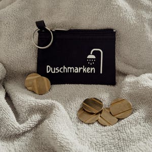 Könnte beinhalten: Eine schwarze Reißverschlusstasche mit silbernem Schlüsselring, dem weißen Text "Duschmarken" und einer Duschkopf-Grafik. Mehrere goldfarbene, runde Marken mit horizontalen Linien liegen um die Tasche herum, alles auf einer grauen, strukturierten Oberfläche.