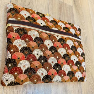 Könnte beinhalten: Rechteckige Clutch mit sich wiederholendem Fächermuster in Braun-, Rot- und Weißtönen. Die Clutch hat einen braunen Reißverschluss und einen goldfarbenen Reißverschlussanhänger. Der Stoff hat ein strukturiertes Aussehen.