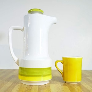 May include: A white ceramic coffee pot with a green lid and yellow and green stripes around the base. A yellow ceramic mug is next to the coffee pot.