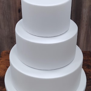 Può includere: Una torta bianca a tre livelli con base rotonda e strati cilindrici. La torta è su una superficie di legno, con uno sfondo di legno. Gli strati della torta diminuiscono di dimensione dal basso verso l'alto.