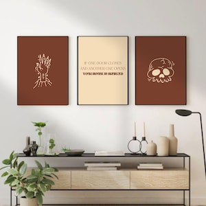 Könnte beinhalten: Drei gerahmte Kunstdrucke. Der linke Druck ist ein brauner Hintergrund mit einer weißen Handillustration. Der mittlere Druck hat den Text: „IF ONE DOOR CLOSES AND ANOTHER ONE OPENS YOUR HOUSE IS INPAINTED“. Der rechte Druck ist ein brauner Hintergrund mit einer weißen Schädelillustration.