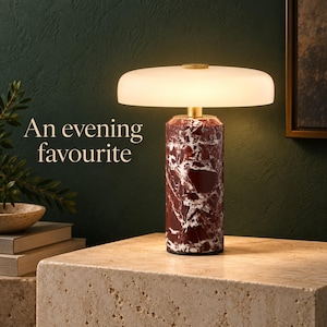 Peut inclure: Une lampe de table avec une base cylindrique en pierre marbrée rouge foncé et blanche, surmontée d'un large abat-jour en verre blanc dépoli lumineux et d'un accent en métal doré. Le texte "An evening favourite" est visible à gauche.