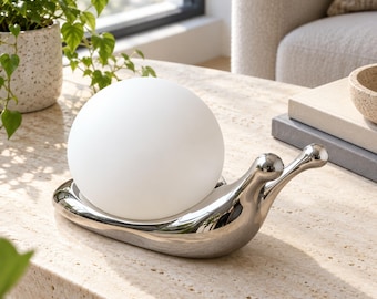 May include: A snail-shaped table lamp featuring a polished silver-tone metallic base and a smooth, spherical white glass shade. The lamp has a curved, elongated body with two rounded antennae extending from the front, supporting the globe light.