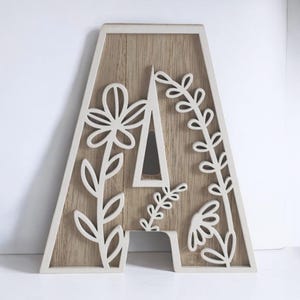 Może przedstawiać: Drewniana litera "A" z jasnobrązowym tłem z usłojeniem drewna i białym obramowaniem. Litera posiada białe kwiatowe wycięcia i dekoracyjny design, odpowiedni do dekoracji wnętrz lub spersonalizowanych prezentów.