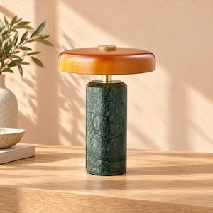 May include: A table lamp featuring a cylindrical dark green marble base with visible veining and a wide, flat, amber-colored glass shade. The lamp is topped with a small, circular, metallic gold-toned knob at the center of the shade.