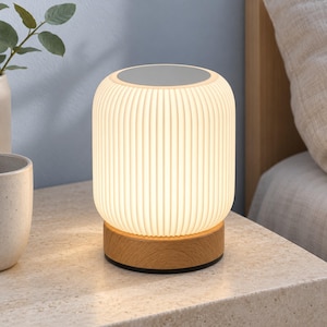 Puede incluir: Una lámpara de mesa cilíndrica con una pantalla blanca translúcida de costillas verticales que emite un brillo suave. La lámpara se apoya sobre una base circular de madera clara con veta visible y una superficie superior plana y sólida.