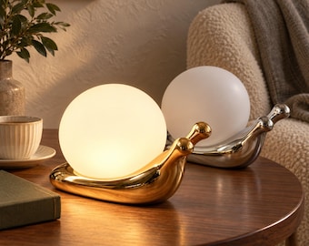 May include: Two snail-shaped table lamps with smooth, metallic bodies and spherical glass shades. One lamp has a polished gold finish with a warm glowing light, while the other has a silver finish with an unlit, opaque white glass globe.
