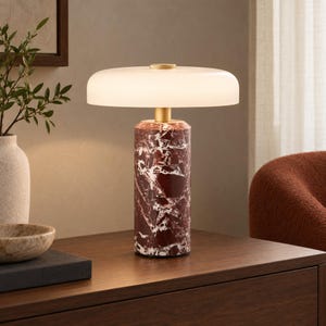 Op de afbeelding: Een tafellamp met een cilindrische voet van donkerrood marmer met witte aders. De lamp heeft een goudkleurige metalen hals en een brede, platte, ronde kap van matglas die een zacht, diffuus licht verspreidt vanuit het midden.