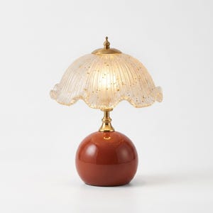 Peut inclure: Une lampe de table avec une base sphérique brillante de couleur terre cuite et un col en métal doré. L'abat-jour en verre cannelé translucide présente un bord festonné, des éclats dorés et un petit fleuron en laiton au sommet.