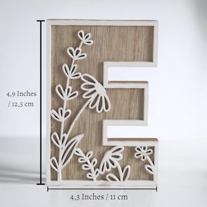 May include: Wooden letter "E" with a floral design. The letter is 12.5 cm tall and 11 cm wide. The letter is white with a light brown wood grain background. White floral cutouts include daisies and other flowers.