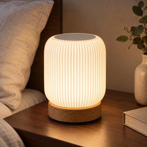 Puede incluir: Una lámpara de mesa cilíndrica con una pantalla blanca de costillas verticales que emite una luz cálida, montada sobre una base circular de madera clara. La lámpara tiene una superficie superior plana y una textura uniforme.