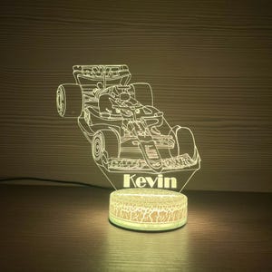 Op de afbeelding: Een oplichtend LED-nachtlampje met een Formule 1-raceauto-ontwerp. Het licht is wit en heeft de naam "Kevin" aan de basis. De basis van het licht heeft een rond, gestructureerd patroon.