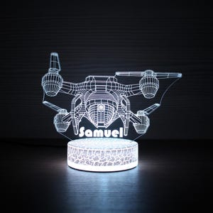 May include: A white, illuminated drone-shaped night light with the name "Samuel" displayed. The light has a detailed wireframe design of a drone, with a circular base. The light is on a dark surface.
