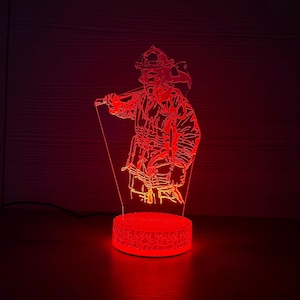 Könnte beinhalten: Rote LED-Lampe, die einen Feuerwehrmann in voller Ausrüstung mit einer Axt zeigt. Die Lampe hat einen runden Sockel und eine detaillierte Umrisszeichnung des Feuerwehrmanns. Das Bild wird von einem roten Schein beleuchtet.