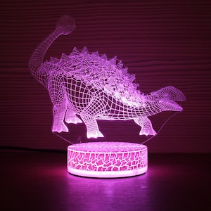 May include: A purple 3D dinosaur night light. The Ankylosaurus is rendered with a grid pattern. The round base has a cracked pattern. The light sits on a dark wooden surface. The overall effect is a glowing, decorative piece.
