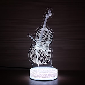 Op de afbeelding: Een witte LED-lamp in de vorm van een cello. Het gedetailleerde draadframeontwerp toont een cello met strijkstok, geplaatst op een ronde, gedecoreerde basis. De lamp straalt zacht licht uit, ideaal voor een muziekkamer. Hoogte: ongeveer 20 cm.