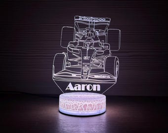 May include: A Formula 1 race car night light with the name "Aaron" etched into the base. The car is white with black outlines, and the base is a light pink color. The light is on, illuminating the car.