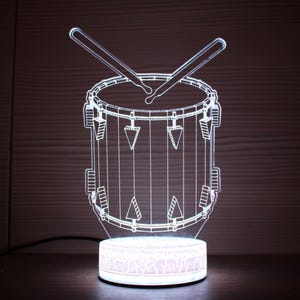Puede incluir: Una luz de noche de batería iluminada en blanco. El diseño presenta un tambor con baquetas encima, apoyado sobre una base redonda. La luz es una ilusión 3D, creando una pieza decorativa única.