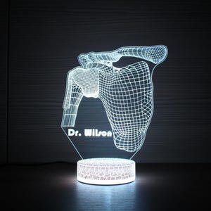 以下が含まれることがあります： 光る3Dレンダリングされた解剖学的肩甲骨ランプ。デザインは、肩甲骨と上腕骨のワイヤーフレームスタイルのレンダリングを特徴とし、ベースには「Dr. Wilson」の文字があります。ランプは丸くテクスチャ加工されたベースにあります。